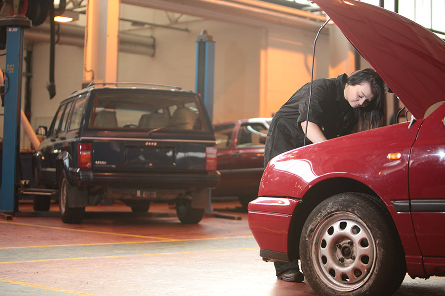Student working under the bonnet of a red car in a garage with other vehicles and equipment visible in the background