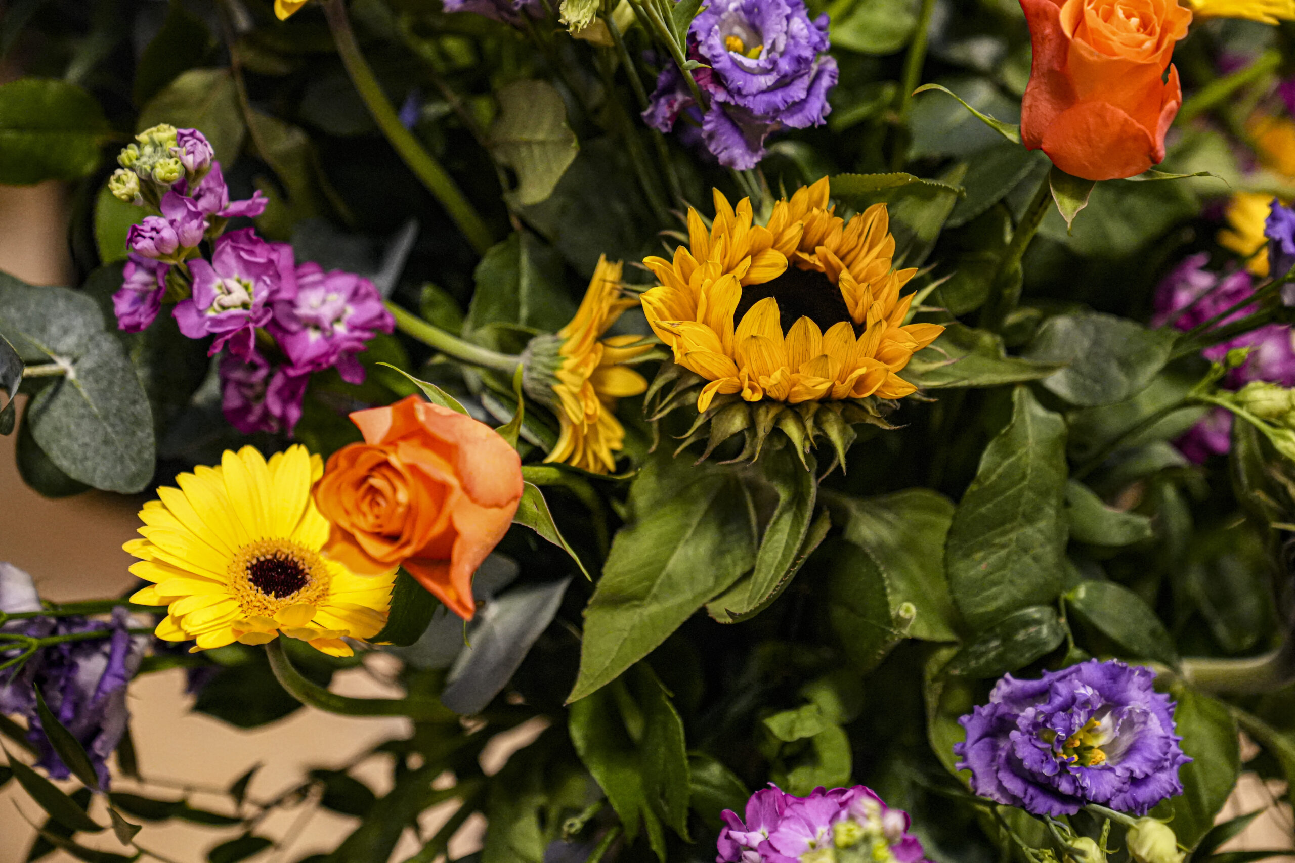 Close-up of a colorful floral arrangement featuring orange roses, yellow gerbera daisies, purple lisianthus, and green foliage.