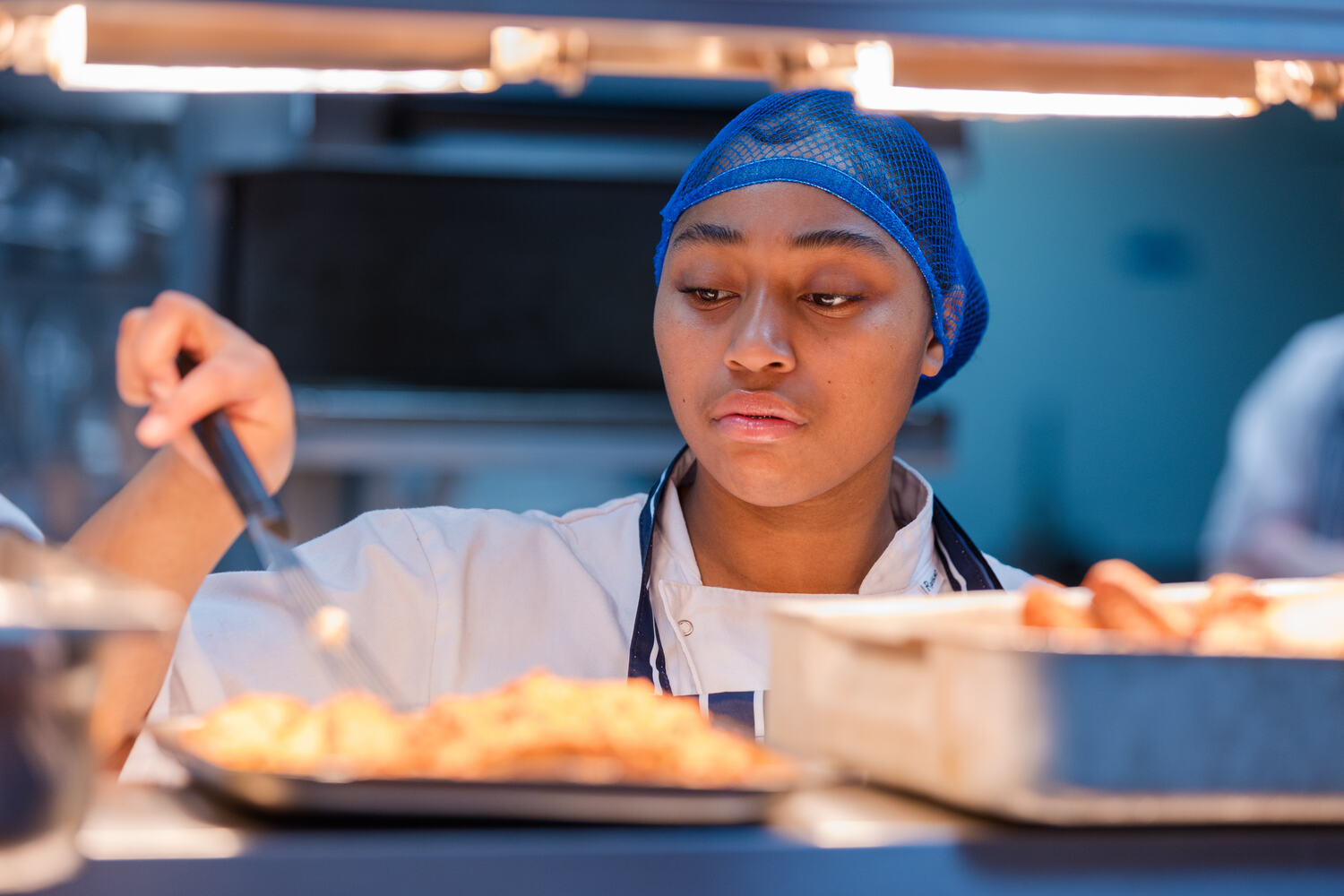 Hospitality and Catering student wearing a chef uniform and blue hairnet serving food from trays in a commercial kitchen setting.