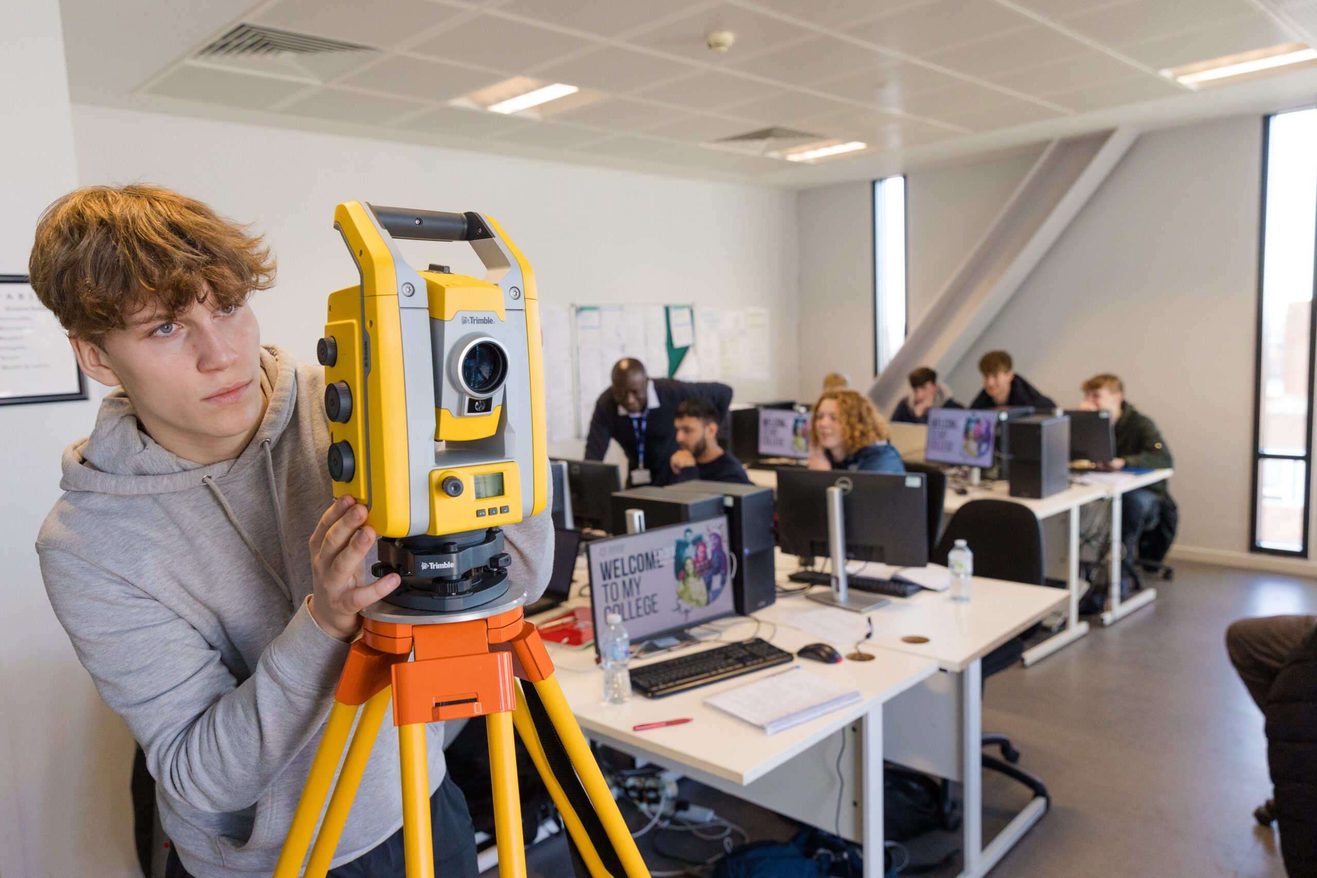 Student operating a yellow tripod-mounted surveying instrument in a classroom with students working on computers in the background.